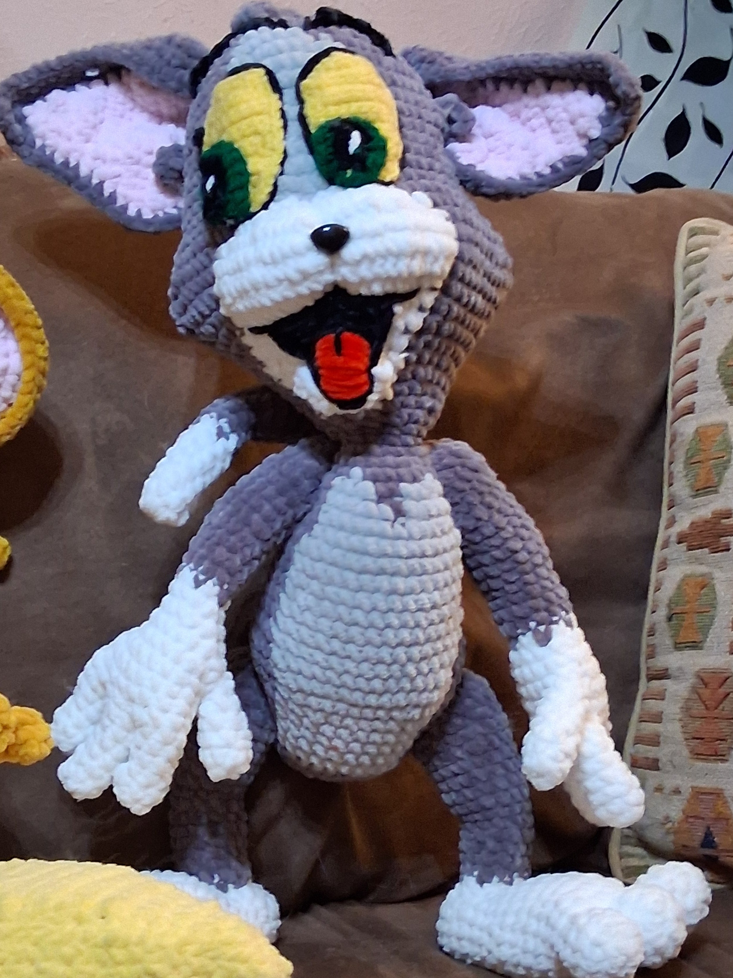 A crocheted plush toy of Tom and Jerry's cat character, featuring gray and white yarn with yellow, green, and red facial details, lying on a brown couch next to a yellow plush toy.